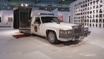 Edward & Nancy Kienholz / 
The Caddy Court, 1986-1987 / 
mobile tableau: 1978 Cadillac with 1966 Dodge van, plaster casts, photographs, wood, metal, cloth, books, paint, polyester resin, light, shelves, curtain, antlers, animal skulls, taxidermy animal heads, clothing, furniture, clock, American flag, microphones, pitcher, glasses, gavel / 
84 x 276 1/2 x 100 in. (213.4 x 702.3 x 254 cm)