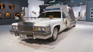 Edward & Nancy Kienholz / 
The Caddy Court, 1986-1987 / 
mobile tableau: 1978 Cadillac with 1966 Dodge van, plaster casts, photographs, wood, metal, cloth, books, paint, polyester resin, light, shelves, curtain, antlers, animal skulls, taxidermy animal heads, clothing, furniture, clock, American flag, microphones, pitcher, glasses, gavel / 
84 x 276 1/2 x 100 in. (213.4 x 702.3 x 254 cm)
