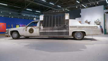 Edward & Nancy Kienholz / 
The Caddy Court, 1986-1987 / 
mobile tableau: 1978 Cadillac with 1966 Dodge van, plaster casts, photographs, wood, metal, cloth, books, paint, polyester resin, light, shelves, curtain, antlers, animal skulls, taxidermy animal heads, clothing, furniture, clock, American flag, microphones, pitcher, glasses, gavel / 
84 x 276 1/2 x 100 in. (213.4 x 702.3 x 254 cm)