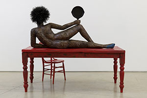 Alison Saar / 
Set to Simmer, 2019 / 
wood, ceiling tin, enamel paint ceiling tin wire, found table, chair, and skillet with texts by Dionne Brand / 
Table and figure: 65 x 72 x 36 in. (165.1 x 182.9 x 91.4 cm) / 
Chair: 38 x 16 x 16 in. (96.5 x 40.6 x 40.6 cm)